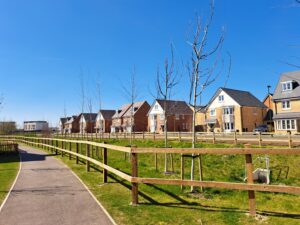Row of modern brick houses along a grassy park with a curved paved walking path and wooden fence under a clear blue sky.