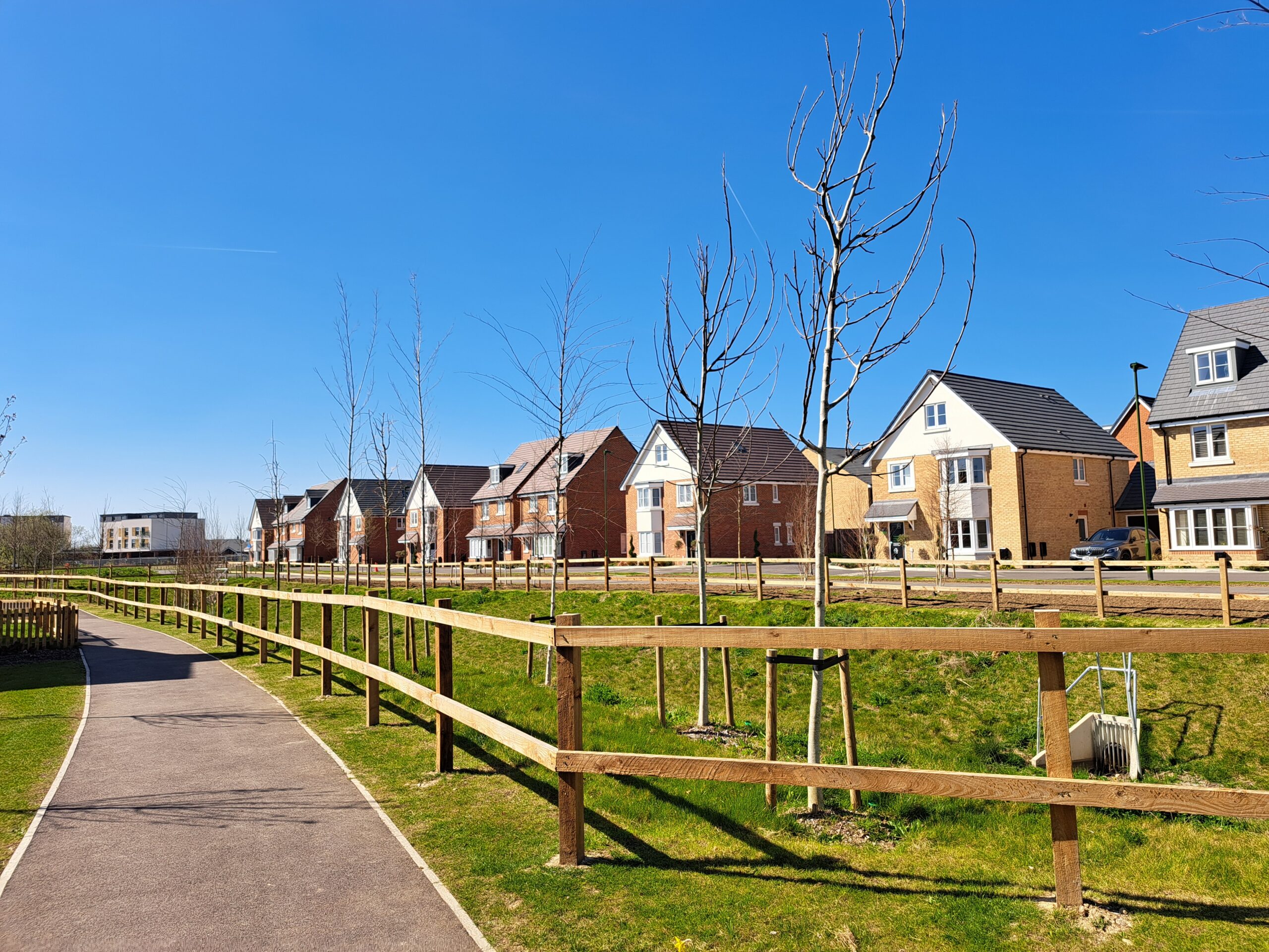Row of modern brick houses along a grassy park with a curved paved walking path and wooden fence under a clear blue sky.