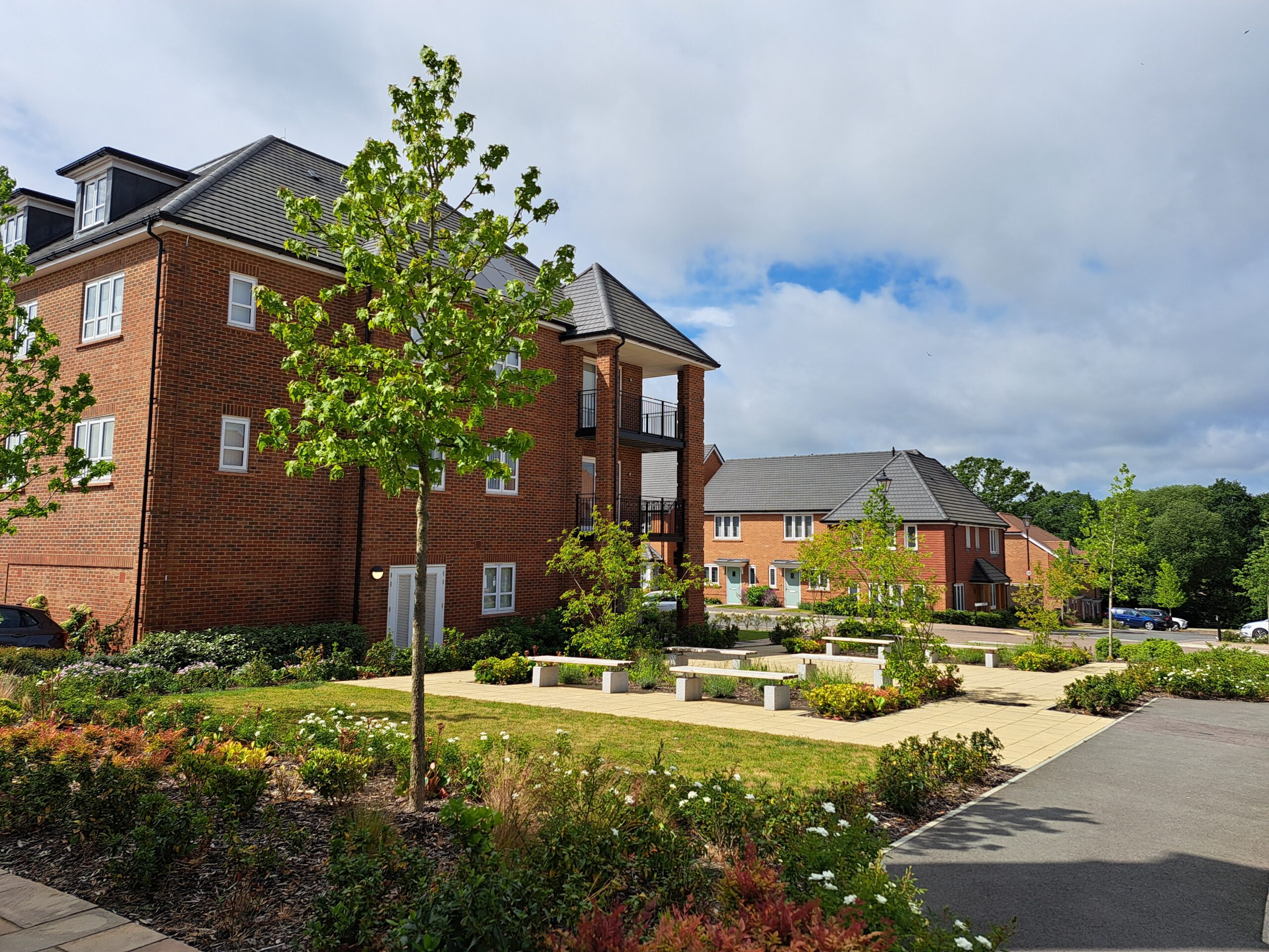 Brick apartment buildings with a landscaped courtyard: trees, shrubs, and concrete benches under a blue sky.