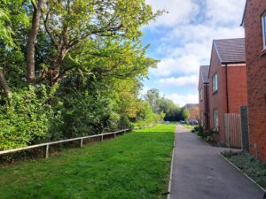 Residential sidewalk flanked by a grassy verge and trees on the left, brick houses on the right under a blue sky with scattered clouds