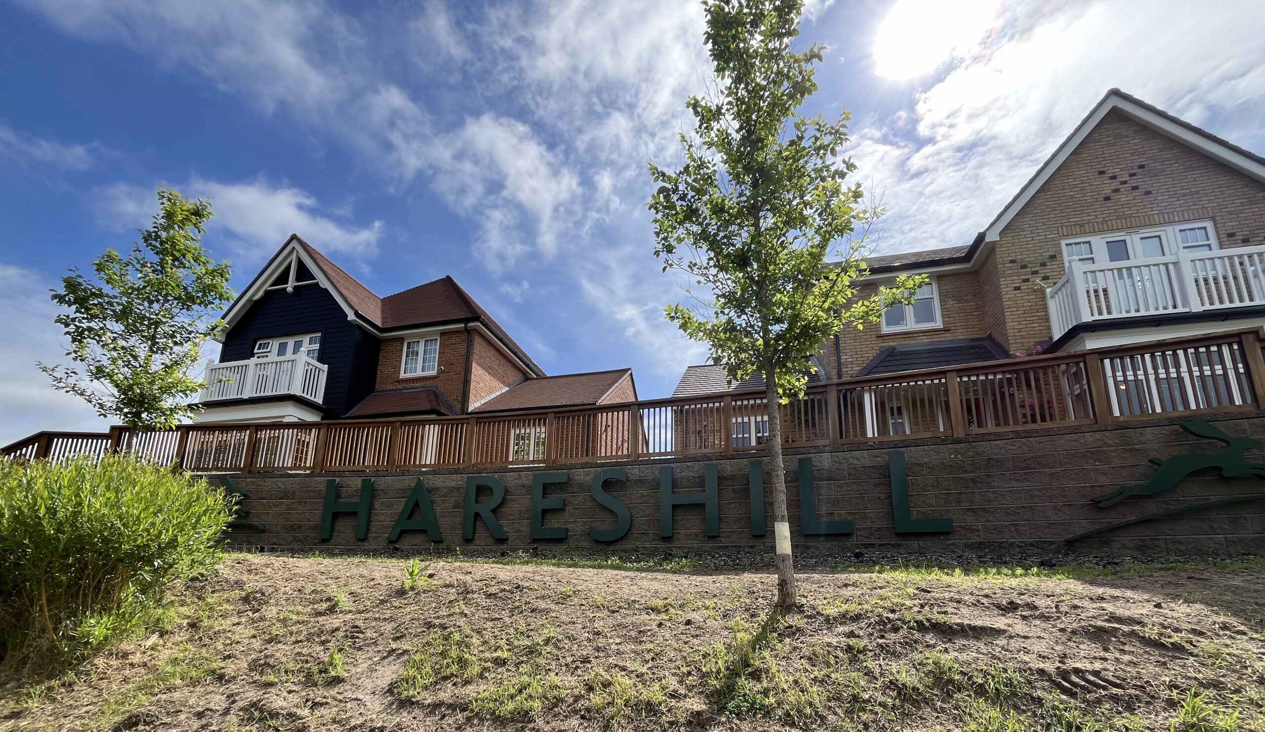 Two-story brick houses behind a wooden railing under a bright blue sky with green trees; large green sign reads 'HARESHILL' along a sloped yard materialled wall.